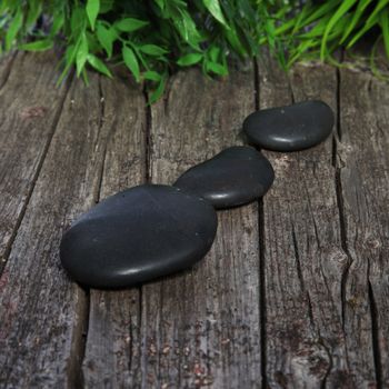 Smooth black basalt spa stones used in hot massage therapy arranged in a row on old rustic weathered wooden boards with greenery in the background