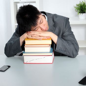 Smart young Asian business man or student sleeping on top of a pile of hardcover books while seated at a table in an office