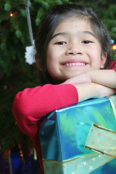 Happy little girl holding her Christmas presents by the Christmas tree. Part Asian, Scandinavian heritage.