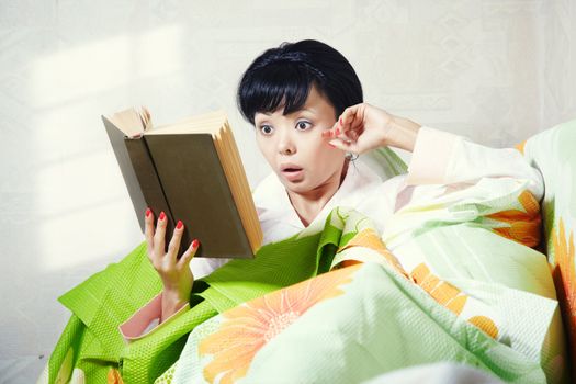 Brunette indoors laying on the bed and reading interesting book