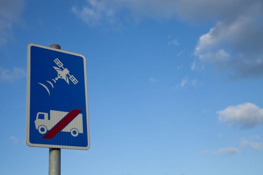 A sign with a lorry, truck, and red slash with a satellite tracking symbol above, on a blue background against a blue sky with cloud