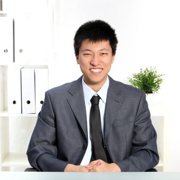 Confident happy young Asian businessman sitting at his desk smiling and facing the camera