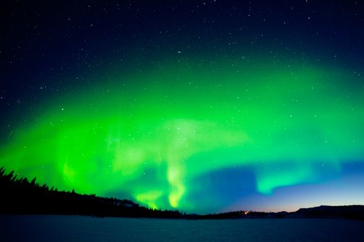 Spectacular display of intense Northern Lights or Aurora borealis forming green swirls over single isolated illuminated house in vast snowy winter landscape of Lake Laberge  Yukon Territory  Canada
