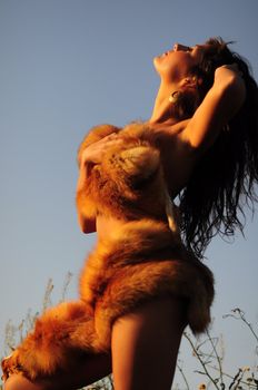 Low angle portrait of a woman wrapped in fox fur standing against the sky in the glow of the setting sun.