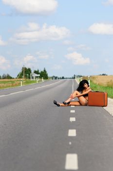 Woman In Bikini On Empty Highway sitting alongside her suitcase with her stiletto shoes in the roadway