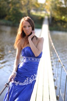 Slender young woman in blue dress standing in the sunshine on a long suspension bridge spanning a river.