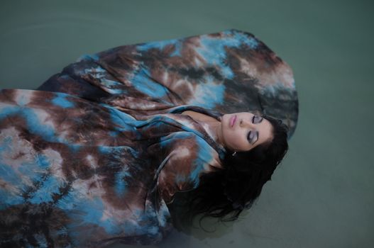 A beautiful brunette woman lies back in the water of a still pond with her colourful dress floating out around her.