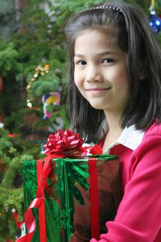 Happy little girl holding her Christmas presents by the Christmas tree. Part Asian, Scandinavian heritage.