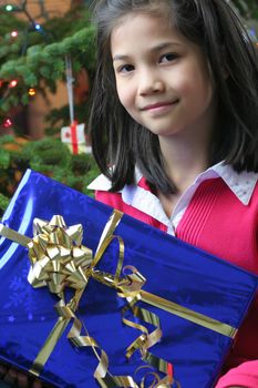 Happy little girl holding her Christmas presents by the Christmas tree. Part Asian, Scandinavian heritage.