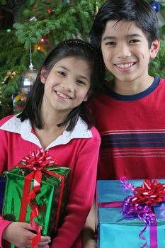 Two happy children holding their Christmas presents by the Christmas tree. Part Asian, Scandinavian heritage.