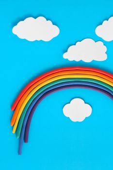 Plasticine rainbow near white paper clouds on blue background