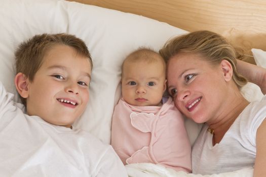 Mother and her kids in the bed, big brother and newborn swaddled sister looking at camera while mother looking at them.