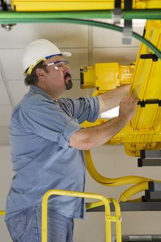 A telecommunication switch worker inspecting Fiberoptic cables in the fiberduct on a ladder platform. He is wearing hardhat and safety glasses. Surrounded by grounding telecom equipment cables.