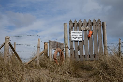 A wooden gate and fence with a 'private' sign and warning 'trespassers will be prosecuted' on a sand dune against a blue sky with cloud.