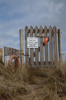 A wooden gate and fence with a 'private' sign and warning 'trespassers will be prosecuted' on a sand dune against a blue sky with cloud.