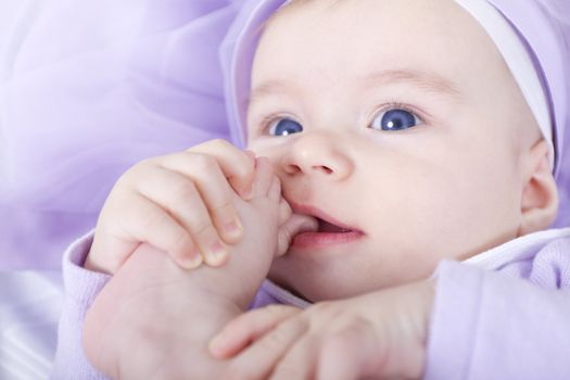 Blue eyed six months old lady holding her toe in her mouth. Wearing Purple Dress and purple background.