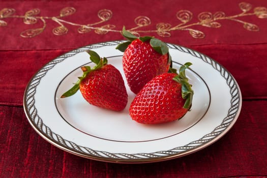 Three red strawberries with green leaves on a white fine china plate with silver decoration on the rim. Red textured background.