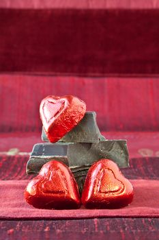 Red foil wrapped candy hearts arranged on top of pile of gourmet thick dark chocolate bar chunks. Red textured background.