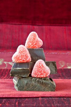 Jelly (gummy) sugar coated candy hearts arranged on top of pile of gourmet thick dark chocolate bar chunks. Red textured background.