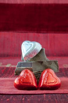 Two red and one silver foil wrapped candy hearts arranged on top of pile of gourmet thick dark chocolate bar chunks. Red textured background. Concept: third wheel, odd man out, does not fit in.