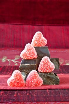 Jelly (gummy) sugar coated candy hearts arranged on top of pile of gourmet thick dark chocolate bar chunks. Red textured background.