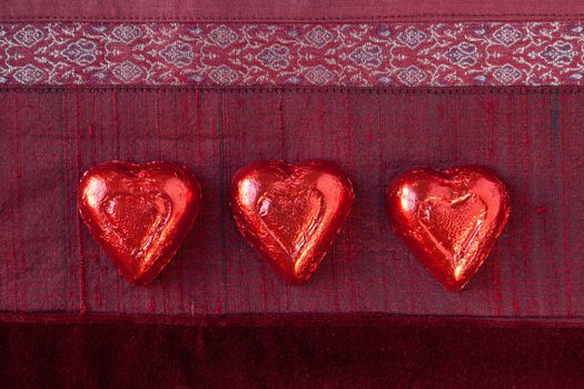 Three red foil wrapped candy hearts in a row on red textured background.