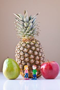 Toy girl and boy standing in front of pineapple, red apple and green pear on white with taupe brown background. The concepts depicted in this image are nutrition, good food choices, balanced diet and good for you.