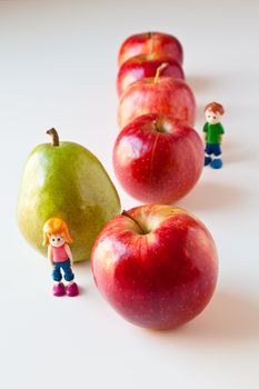 Toy girl and boy overwhelmed by making good food choices. Green pear standing out from a line of red apples. The concepts depicted in this image are nutrition, good food choices, balanced diet, good for you, being different, unique, stick out, being singled out.