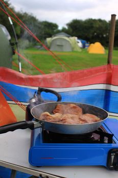 A frying pan on a small blue camping gas stove with frying bacon. All set in a campsite setting with tents in the background, and a brightly coloured wind break for protection.