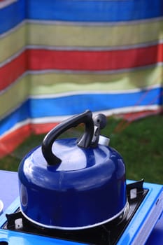 A blue camping kettle on a gas stove with a brightly coloured wind breaker for protection against the wind in soft focus to the background. Set on a portrait format.