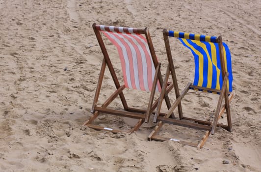 Two wooden deckchairs on the beach. Both covered in stripped nylon in red and white and yellow and blue.