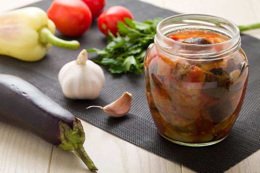 Cooked stewed eggplants in jar for canning with raw vegetables:  garlic, parsley, tomatoes, pepper on wooden table