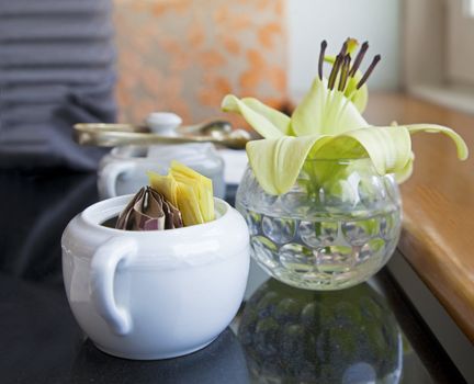 Interior corner detail of black top table with tea and coffee utensils of sugar bowls and bud vase with a lily by a window