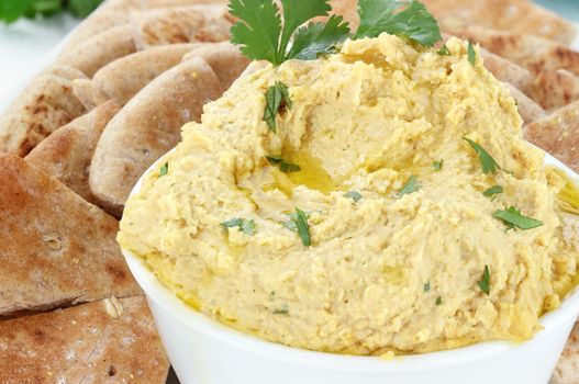 Bowl of hummus topped with pure olive oil and cilantro and served with wedges of pita bread for a healthy snack.
