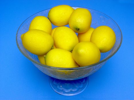 A decorative bowl, engraved with floral patterns, full of lemons, viewed from about 45 degrees above and placed over a mid-blue background.