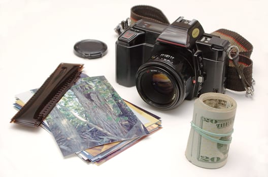 An SLR camera sits alongside a pile of photos and strips of 35mm negatives, with a roll of  bills standing beside them, on a white background.