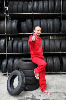 A positive mechanic in a garage standing next to a rack full of tires and making a positive gesture.