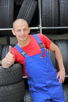 A smiling mechanic in a garage standing next to a rack full of tires and making a positive gesture.