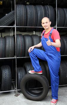 A smiling mechanic in a garage standing next to a rack full of tires and making a positive gesture.