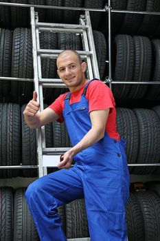 A smiling mechanic in a garage standing on a ladder next to a rack full of tires and making a positive gesture.