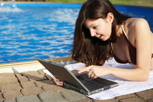 A very attractive young woman sunbathing next to a swimming pool and writing digital holiday greetings on her notebook computer.