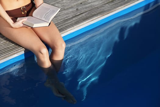 An attractive young woman wearing a bikini and reading a book next to the swimming pool