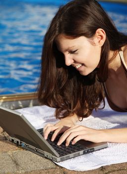 A very attractive young woman sunbathing next to a swimming pool and writing digital holiday greetings on her notebook computer.