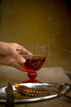A hand holding a glass with red wine and a plate with barbecued octopus piece