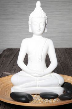 White statue of a meditating buddha sitting in an earthenware bowl with smooth black basalt massage stones in a spa for tranquillity and relaxation