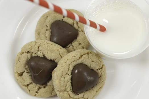 three cookies with chocolate hearts and glass of milk with a striped red straw view from above with white background