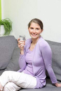 Happy healthy mature woman sitting on a sofa in a living room drinking a glass of fresh pure water looking at the camera with a smile