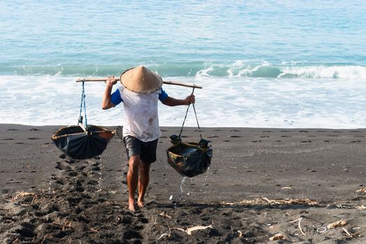 Manual male worker collects water for sea salt production on volcanic black sand in Amuk Bay, Bali, Indonesia. Salt farmers follow a unique tradition of salt production dating back over 900 years. 
