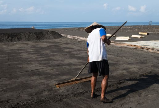 Manual male worker collects black volcanic sand for sea salt production in Amuk Bay, Bali, Indonesia. Salt farmers follow a unique tradition of production dating back over 900 years. 