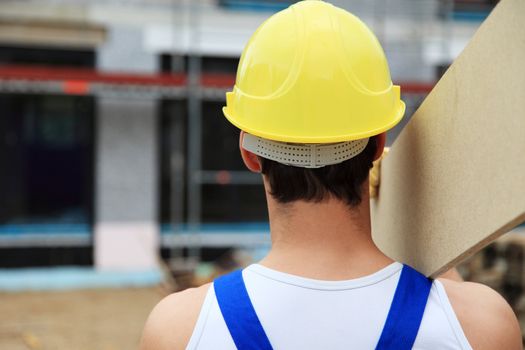 Backview of a manual worker on construction site carrying wooden board.